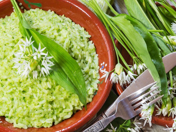 Grünes Bärlauch-Risotto in einem rötlichen Teller aus Ton neben frischen Bärlauch-Blättern auf einem hellen Holztisch mit Besteck von oben.