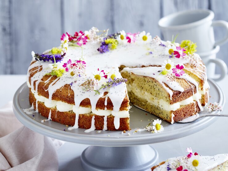 Zitronen-Drizzle-Kuchen mit Mohn / Deko aus frischen Blüten / auf Kuchenplatte angerichtet / Hintergrund: zwei Tassen