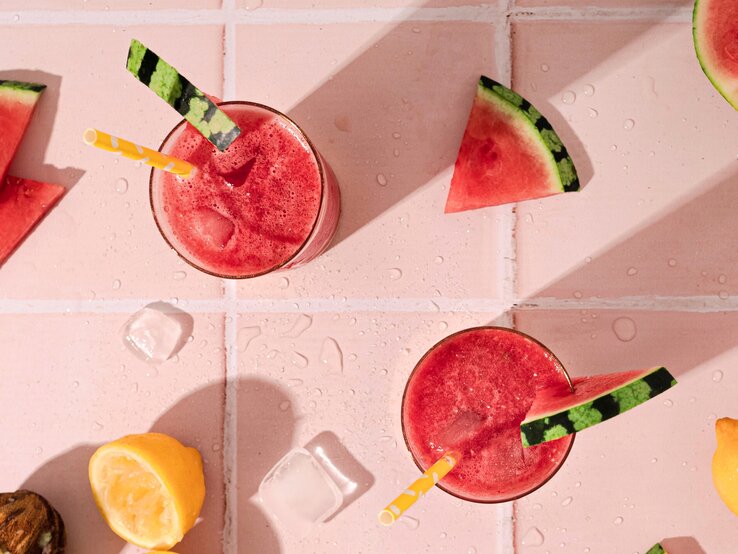 Dieser sommerliche Cocktail ist die pure Erfrischung und kühlt dich an jedem noch so heißen Tag – Wassermelonen-Frosé. Wassermelone Frosé in zwei großen Gläsern mit Strohhalm und frischer Melone auf einem rosa Tisch aus Fliesen.