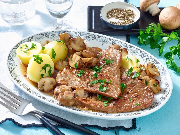 Schnitzel mit Champignons und Kartoffeln / ovaler Teller aus Porzellen/ Gabel / frische Petersilie / kleine runde Schale mit Pfefferkörnern / zwei Gläser / zwei frische Champignons / Draufsicht, schräg