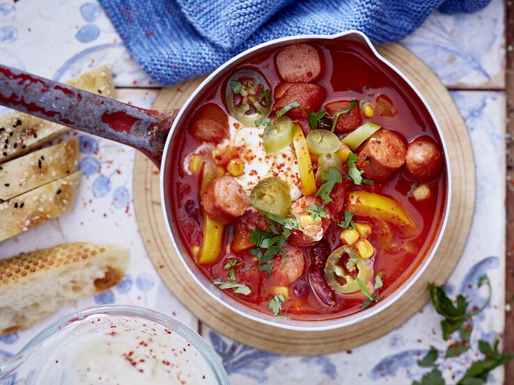 mexikanisches Würstchen-Gulasch Ein Topf mit kräftig roter Suppe, gefüllt mit geschnittenen Würstchen, Mais, Kidneybohnen, Paprika, Jalapeños und einem Klecks Sauerrahm, garniert mit frischen Kräutern. Daneben liegen aufgeschnittenes Fladenbrot und ein Glas mit einer hellen Sauce. Der Topf steht auf einem runden Holzbrett auf einem gemusterten Untergrund.