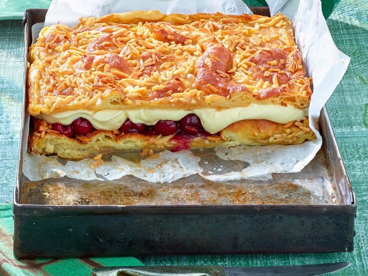 Das Bild zeigt eine Backform mit einem rechteckigen Kuchen, bestehend aus mehreren Schichten. Die oberste Schicht ist mit gerösteten Mandelsplittern bedeckt, darunter befindet sich eine Schicht aus cremiger Füllung und eine weitere Schicht mit eingelegten Früchten, vermutlich Kirschen. Darunter liegt eine Schicht aus Teig, die ebenfalls goldbraun gebacken ist.