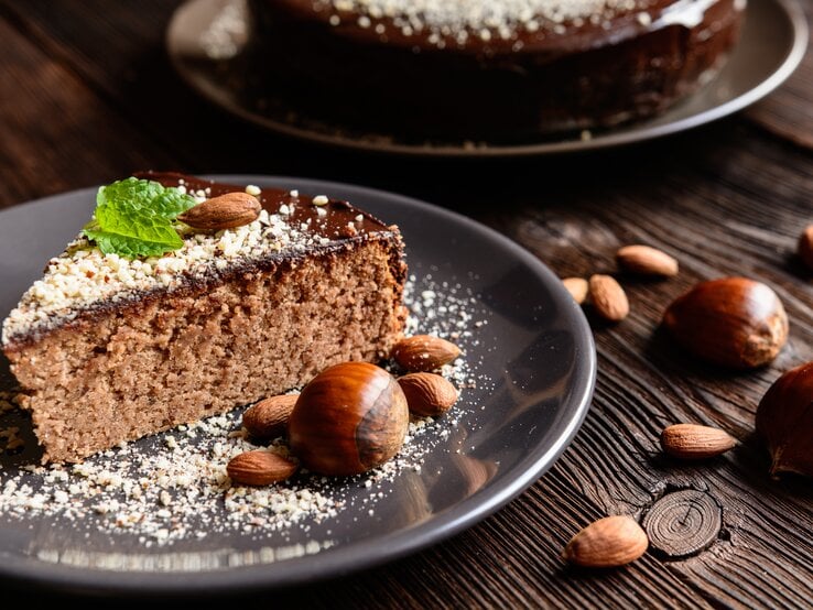 Ein angeschnittenes Stück Maronenkuchen auf einem schwarzen Teller. Dieser steht auf einem dunklen Holztisch.