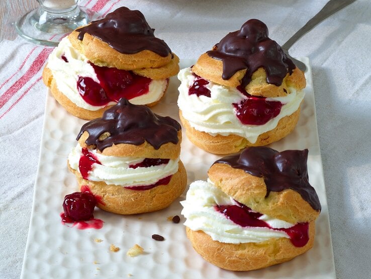 Vier Windbeutel mit Sahne, Kirschfüllung und glänzendem Schokoladenguss liegen auf einer weißen rechteckigen Platte.