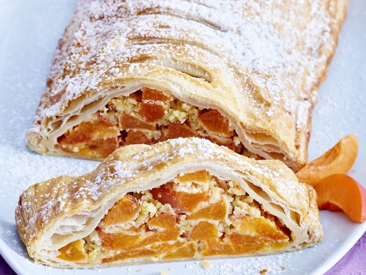 Aprikosen-Mandel-Strudel mit Puderzucker bestreut und angeschnitten serviert auf einem weißen Teller auf rosanem Untergrund. 