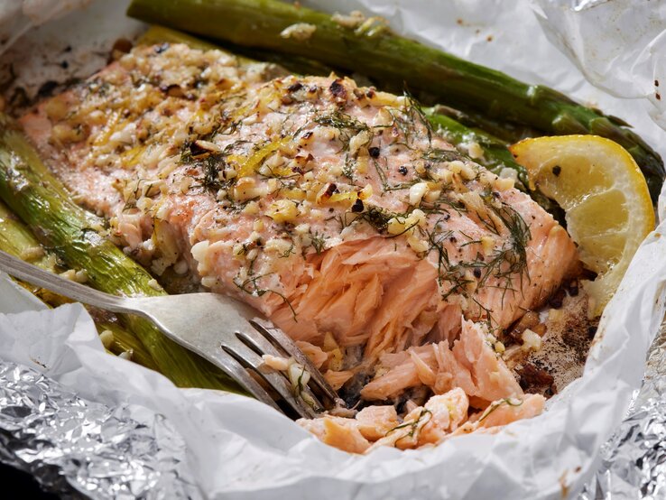 Ofenlachs mit grünem Spargel, Zitrone, Knoblauch und Dill in Backpapier und Alufolie mit einer Gabel. Draufsicht.
