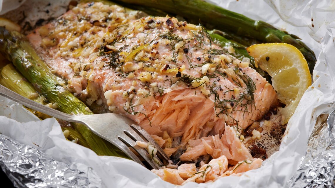 Ofenlachs mit grünem Spargel, Zitrone, Knoblauch und Dill in Backpapier und Alufolie mit einer Gabel. Draufsicht.