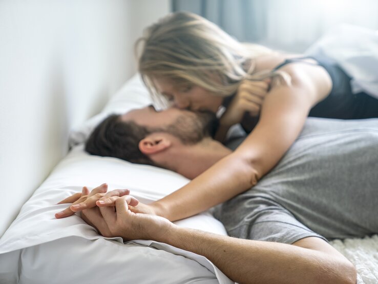 Junges Paar liegt küssend und Händchen haltend im Bett. 