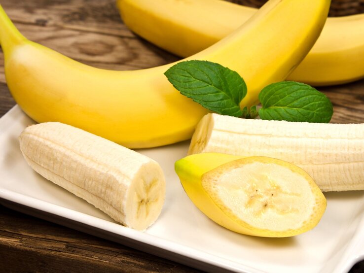 Bananen auf einem Tisch | © Adobe Stock / PhotoSG Frische Bananen, teils geschält und geschnitten, liegen dekorativ mit Minzblättern auf einem weißen Teller. | © Adobe Stock / PhotoSG