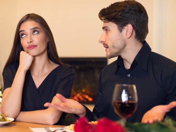 junge Frau sitzt mit ihrem Partner an einem Tisch, er redet auf sie ein, sie rollt die Augen genervt nach oben.