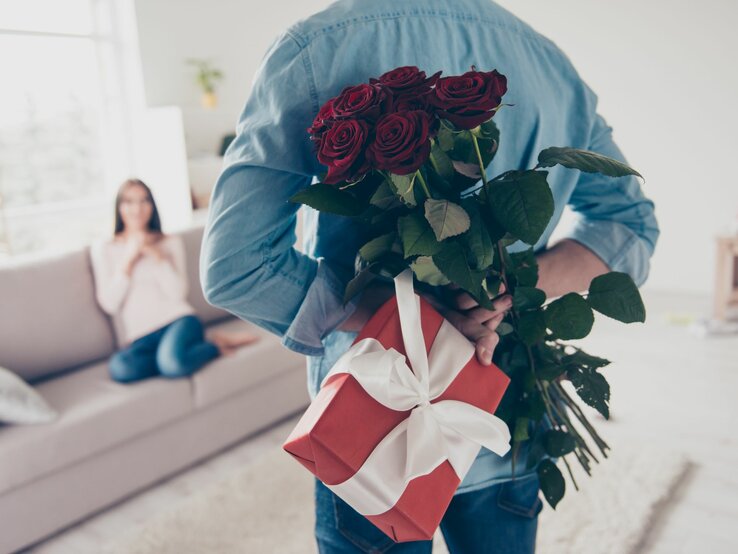 Junger Mann von hinten, der hinter seinem Rücken Rosen und ein Geschenk hält. Vor ihm ist schemenhaft eine Frau auf einem Sofa zu erkennen.