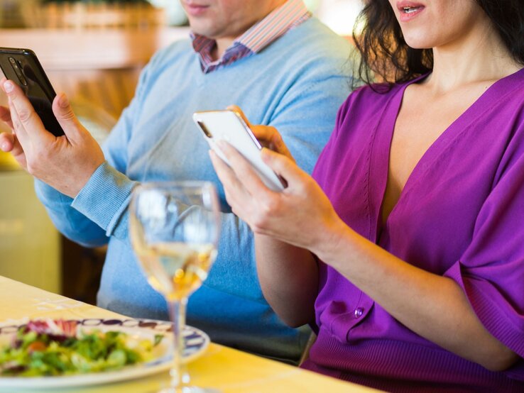 Phubbing Eine Frau in lila Kleid und ein Mann in blauem Pulli starren beim Essen nebeneinander auf ihre Smartphones, ohne sich anzuschauen.