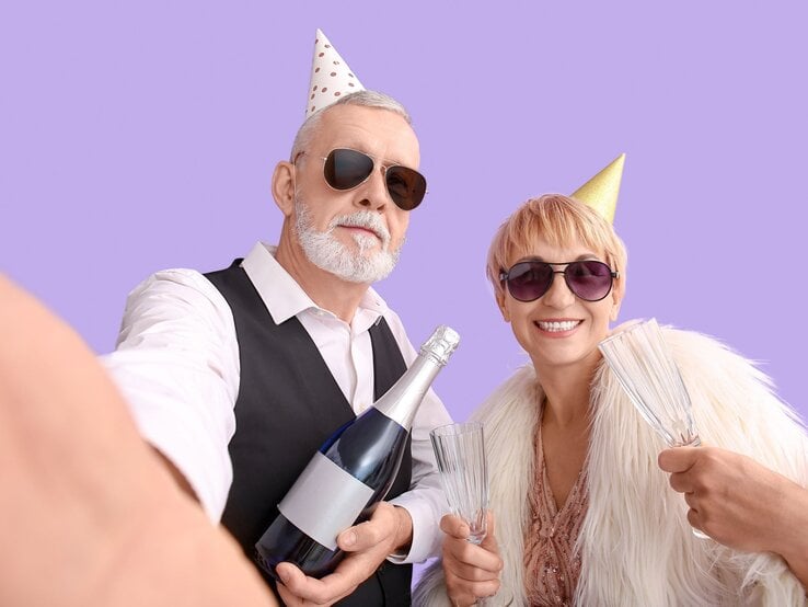 älteres Paar nimmt Selfie von sich auf Älteres Paar mit Sonnenbrillen und Partyhüten macht fröhlich ein Selfie mit Sektflasche und Gläsern vor lila Hintergrund.