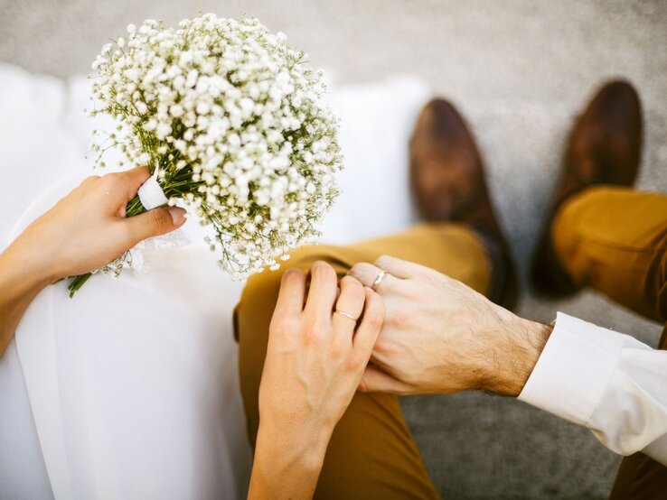 Ein Brautpaar sitzt nah beieinander, hält sich zärtlich an den Händen, die Braut trägt Schleier und ein Sträußchen Schleierkraut.