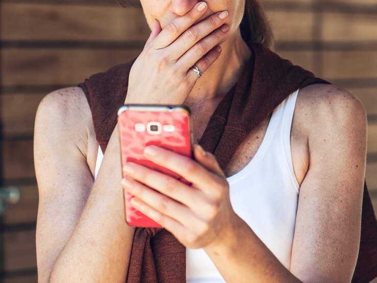 Eine Frau mit heller Haut und blondem Haar in weißem Top und braunem Pullover blickt nachdenklich auf ein pinkes Smartphone vor Holzwand.