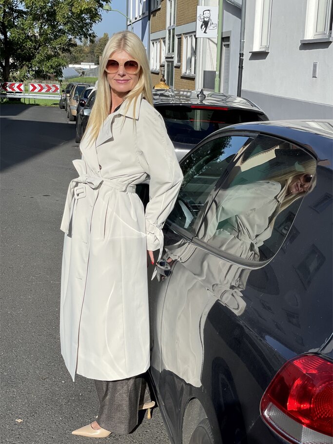Frau in langem weißen Trenchcoat und beigefarbenen Schuhen lehnt seitlich an einem schwarzen Auto, sonnige Straße im Hintergrund. | © Renate Zott