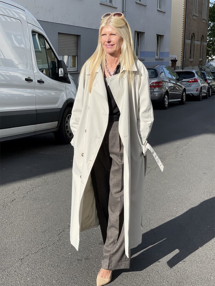 Frau mit langen blonden Haaren in weißem Trenchcoat, schwarzem Oberteil, grauer Hose und beigen Absatzschuhen auf sonniger Straße. | © Renate Zott