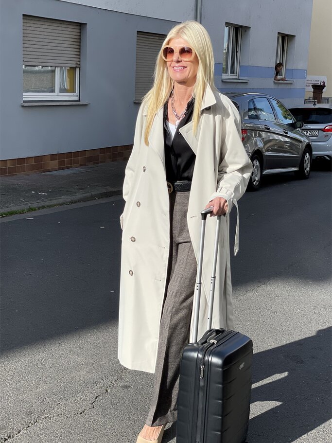 Frau in weißem Trenchcoat, schwarzem Oberteil und grauer Hose zieht einen schwarzen Rollkoffer auf einer Wohnstraße. | © Renate Zott