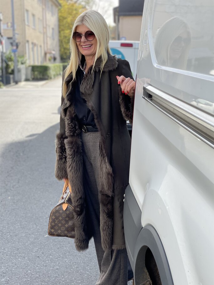 Frau in schwarzer Bluse, braunem Mantel mit Fellbesatz und grauer Hose lehnt an einem weißen Fahrzeug und hält eine Designer-Handtasche. | © Renate Zott