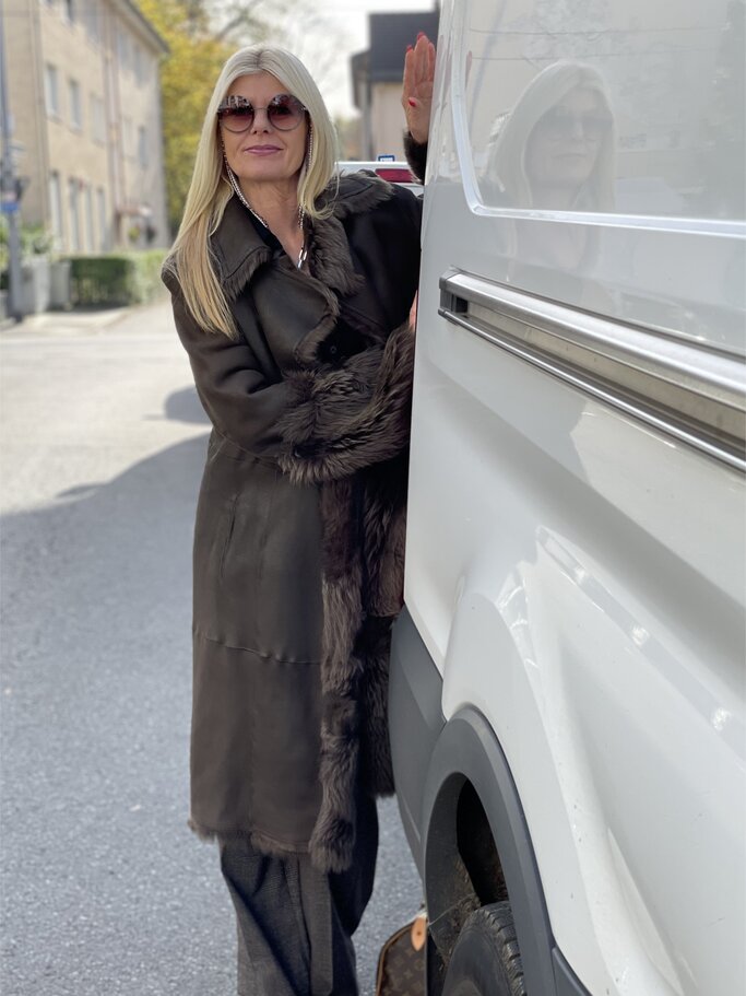 Frau in langem braunem Mantel mit Fellbesatz trägt graue Hose und beigefarbene Schuhe, lehnt an einem weißen Lieferwagen. | © Renate Zott