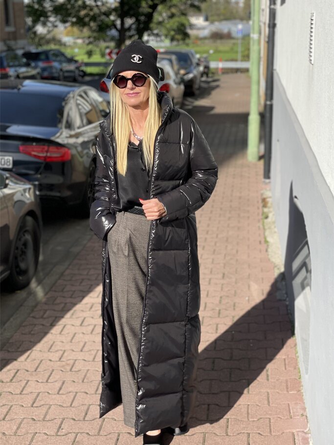 Frau in langem schwarzen Daunenmantel, grauer Hose und schwarzen Schuhen steht auf gepflastertem Weg, parkende Autos im Hintergrund. | © Renate Zott