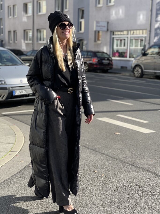 Frau in langem schwarzen Daunenmantel, grauer Hose und schwarzen Schuhen steht auf gepflastertem Weg, parkende Autos im Hintergrund. | © Renate Zott