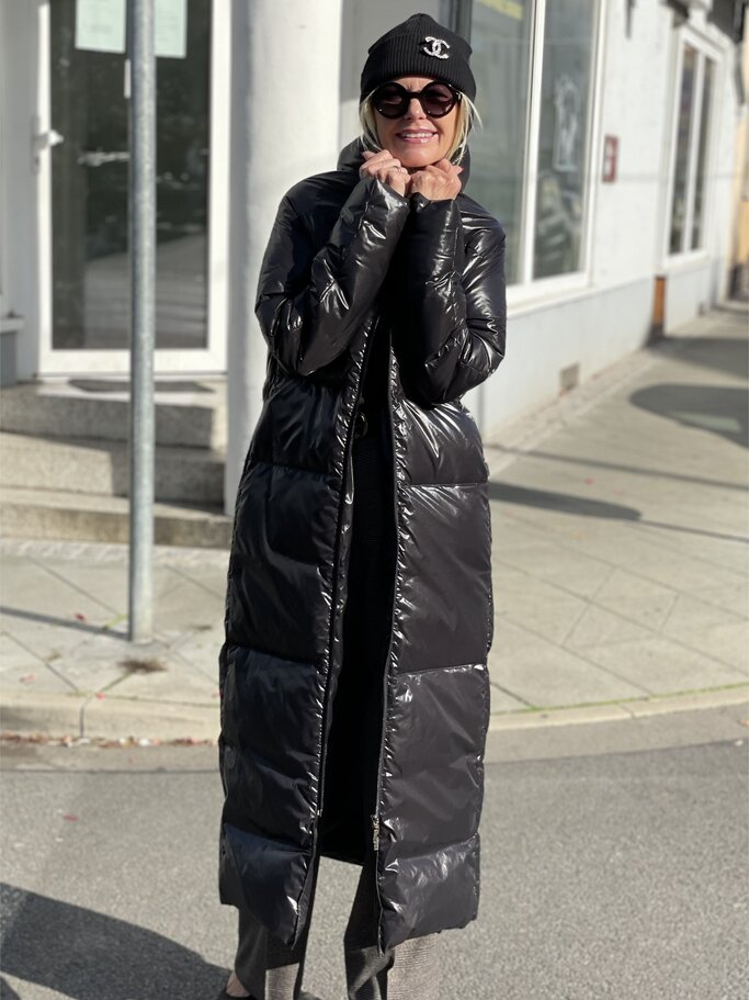 Frau in langem schwarzen Daunenmantel, grauer Hose und schwarzen Schuhen steht auf gepflastertem Weg, parkende Autos im Hintergrund. | © Renate Zott