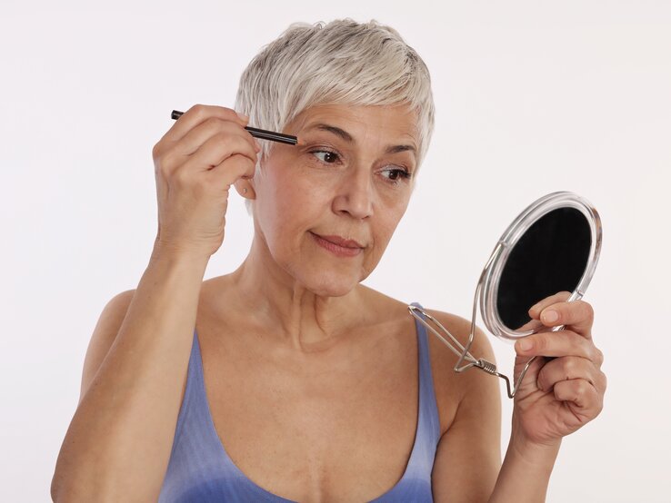 Eine ältere Frau mit silbergrauem Haar trägt Makeup auf, während sie in einen Handspiegel schaut. Sie benutzt einen Lidschattenstift und hat einen konzentrierten Blick. Die Frau trägt ein blaues Top und der Hintergrund ist einheitlich weiß, was den Fokus auf ihre Handlung legt.