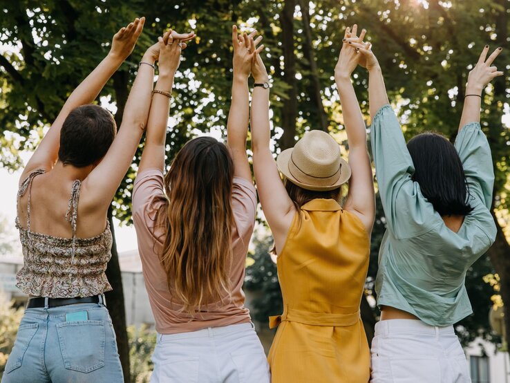 Vier junge Frauen stehen Rücken an Rücken mit erhobenen Armen im Sommeroutfit unter grünen Bäumen in fröhlicher Stimmung. | © Adobe Stock / Bostan Natalia