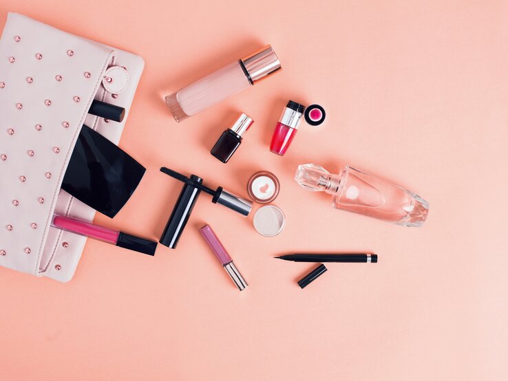 Ein flach angeordnetes Arrangement von Kosmetikprodukten auf einem pastellrosa Hintergrund. Zu den sichtbaren Gegenständen gehören eine hellrosa Kosmetiktasche mit Nieten, mehrere Lippenstifte und Lipglosse in verschiedenen Schattierungen, eine Wimperntusche, ein Flakon mit klarem Parfüm, ein Puderbehälter, ein flüssiges Make-up in einer Glasflasche, ein Nagellack und ein Augenbrauenstift. Die Anordnung ist sorgfältig platziert, um eine ästhetische Komposition zu schaffen, die sowohl Stil als auch die Vielfalt von Beauty-Produkten zeigt. | © iStock/AniDimi