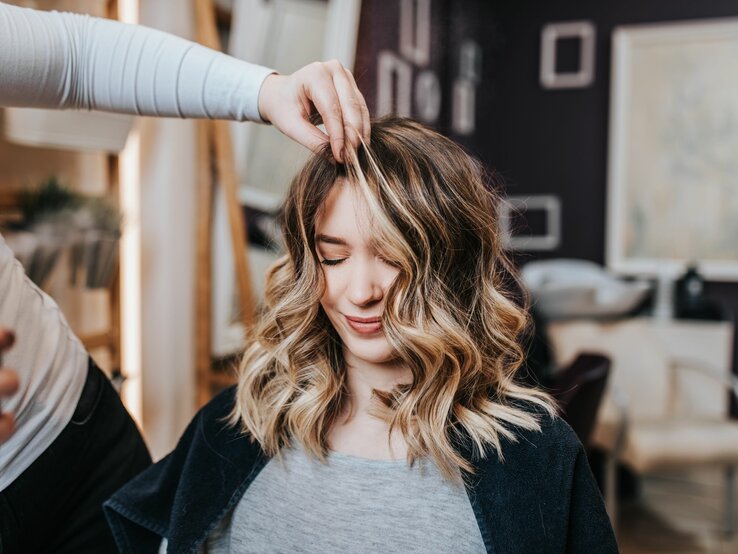 Junge Frau mit mittellangen, gewellten Haaren, die in einem Frisörsalon sitzt. Ihre Haare sind sorgfältig gestylt und weisen blonde Strähnen auf, die einen schönen Kontrast zu den dunkleren Haarpartien bilden. Eine Person, die nur teilweise und unscharf im Hintergrund zu erkennen ist, hebt mit den Fingern eine Strähne ihrer Haare an, vermutlich um die Frisur zu perfektionieren 