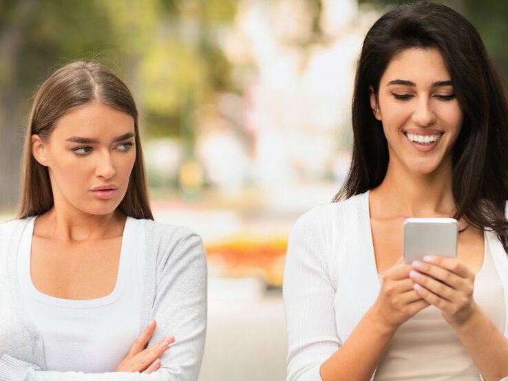 Zwei junge Frauen nebeneinander – die eine ist am Handy, die andere schaut missbilligend drein | © über Spoton