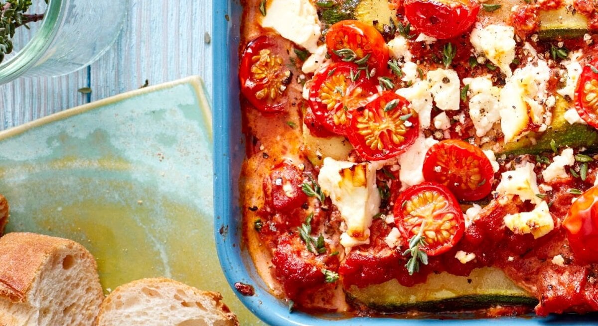 Eine Zucchini-Lasagne mit Feta in einer blauen Auflaufform steht auf einem Geschirrtuch auf einem blauen Tisch. Die Lasagne ist angerichtet mit Feta, Tomaten und Thymian. Neben der Auflaufform liegt Baguette und ein Glas mit Kräutern.