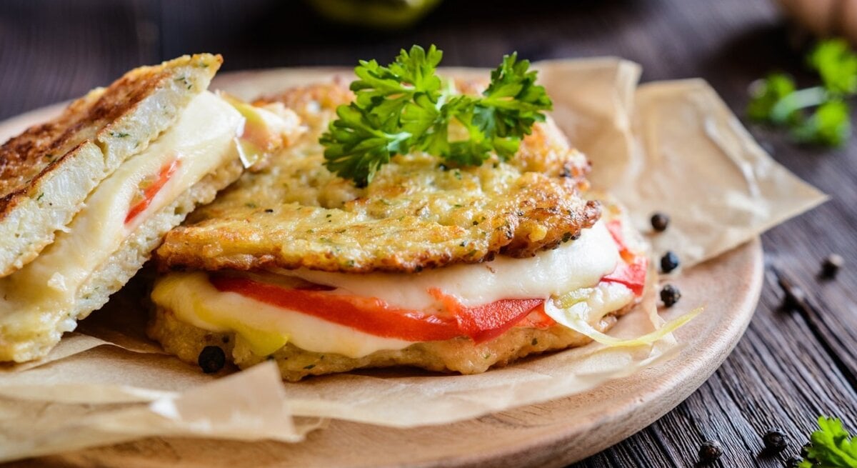Die Low-Carb-Panini mit Mozzarella und Tomate auf einem Brett. Dahinter verschiedene Zutaten auf einem hölzernen Untergrund.