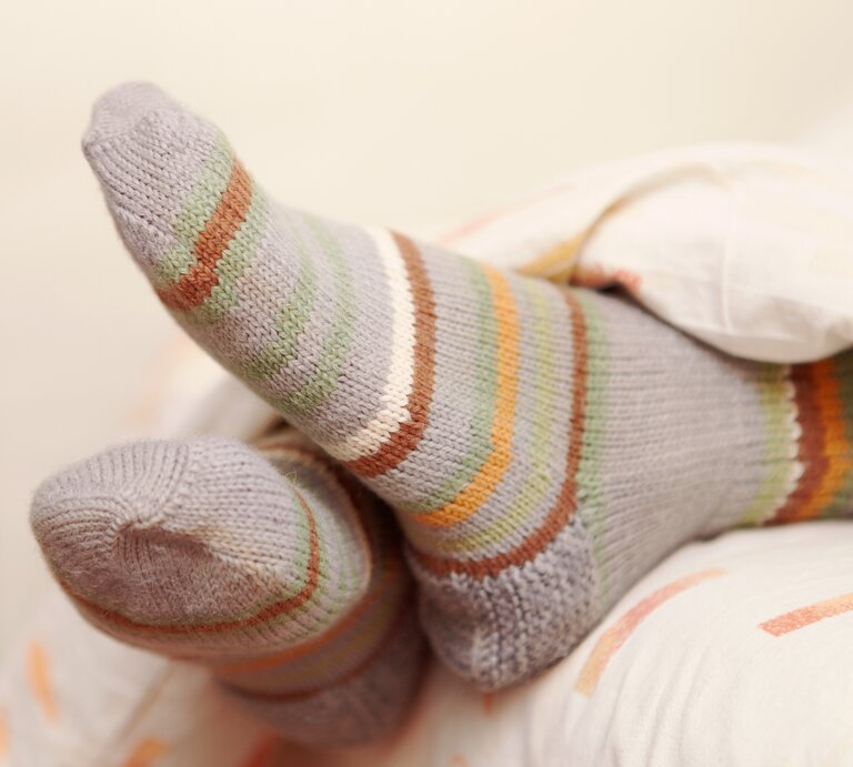 Eine Person liegt entspannt im Bett mit den Füßen hoch, die in gestreiften Socken in sanften Grau-, Grün-, Orange- und Weißtönen gehüllt sind. Der Fokus liegt auf den gemütlichen Socken, die gegen die weiche Bettwäsche drücken. | © Getty Images/Alex_Doubovitsky