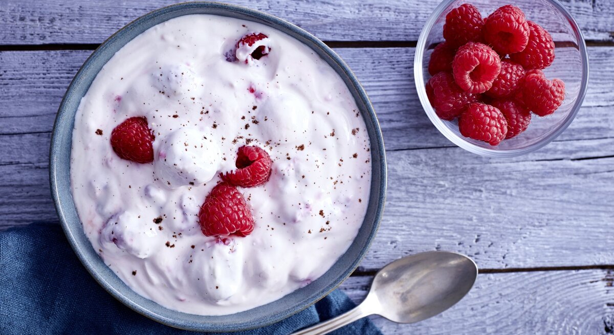 Himbeer-Traum in einer Schale, bestehend aus  Zitronenabrieb, Himbeeren, Honig und griechischem Joghurt, serviert auf einem hölzernen Frühstückstisch. 
