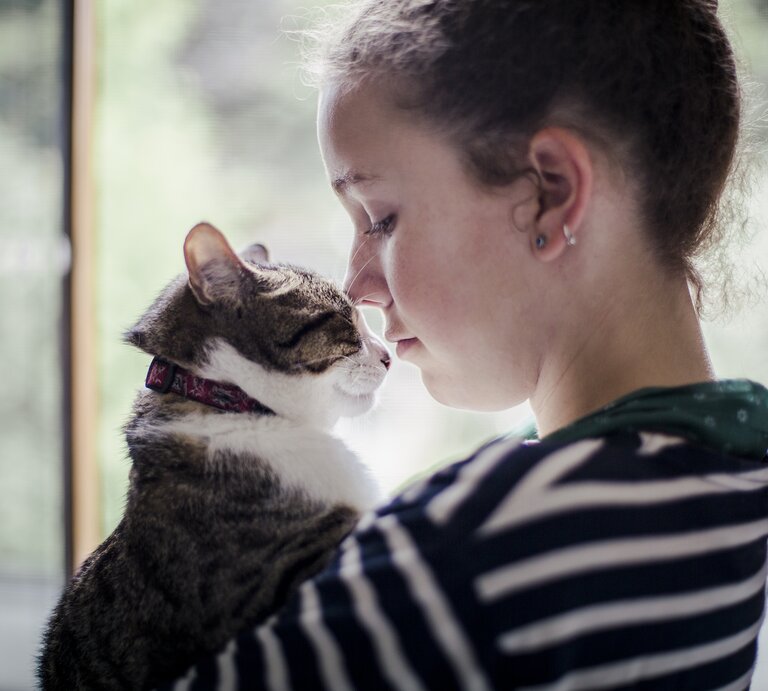 Eine junge Frau steht am Fenster und hält ihre Katze im Arm. Sie schauen sich an.  | © GettyImages/Linda Raymond
