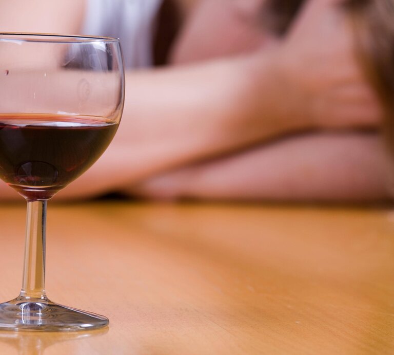 Ein Glas Rotwein auf dem Tisch, im Hintergrund eine verschwommene Person mit langen Haaren. | © IMAGO / Panthermedia