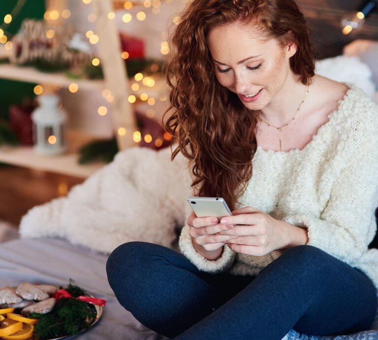 Junge Frau sitzt auf dem Sofa und verschickt Advent Sprüche an ihre Freunde und die Familie. Ihre Wohnung ist weihnachtlich dekoriert.  | © AdobeStock/gpointstudio