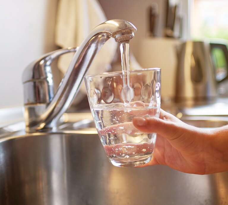 Das Bild zeigt eine Person, die ein Glas mit klarem Wasser aus einem modernen, metallischen Wasserhahn in einer Küchenspüle füllt. Das Tageslicht fällt durch ein Fenster ein und beleuchtet die Szene, was auf eine häusliche und gesunde Umgebung hinweist. Das Wasser sieht sauber aus und die Szene kommuniziert die Zugänglichkeit von sauberem Trinkwasser, was für eine gesunde Lebensweise unerlässlich ist. | © iStock/sonsam