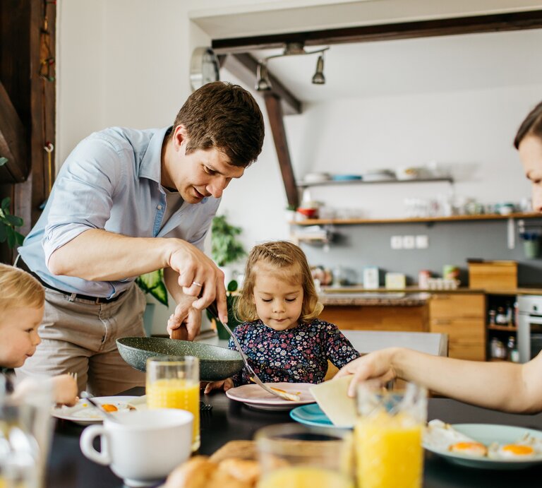 Eine glückliche Familie beim Frühstück: Ein Mann in einem hellblauen Hemd beugt sich über, um seiner kleinen Tochter zu helfen, die konzentriert auf ihren Teller blickt. Ein weiteres Kleinkind sitzt daneben und ist auf sein Essen fokussiert. Eine Frau sitzt gegenüber, reicht etwas hinüber und lächelt dabei. Der Tisch ist mit einem gesunden Frühstück mit Orangensaft, Spiegeleiern und Brötchen gedeckt. Sie befinden sich in einer hellen Küche mit moderner Einrichtung und Pflanzen, die für eine wohnliche Atmosphäre sorgen. | © iStock.com/AnVr