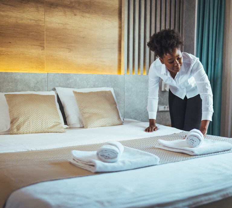 Eine Hotelangestellte streicht den Bezug eine Bettes glatt. | © shutterstock/Dragana Gordic