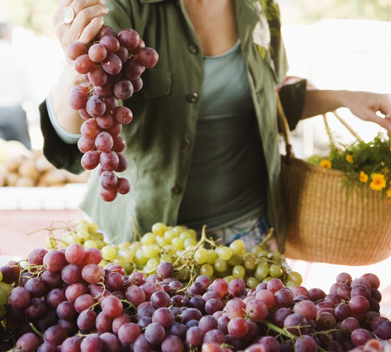Das Bild zeigt eine Frau, die eine Traube roter Trauben über einem Marktstand hält, der mit verschiedenen Traubensorten gefüllt ist. Die Trauben erscheinen frisch und saftig, mit einem Farbspektrum von hellgrün bis dunkellila. Die Frau trägt ein grünes Oberteil und hat einen geflochtenen Korb über dem Arm, der mit Blumen geschmückt ist, was darauf hindeutet, dass sie frische Produkte und Blumen einkauft. Der Fokus liegt auf den Trauben und den Händen der Person, während der Hintergrund mit anderen Marktaktivitäten unscharf bleibt, was eine lebendige Marktatmosphäre vermittelt.  | © GettyImages / Jupiterimages