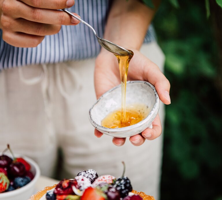 Eine Person gießt mit einem Löffel Reissirup aus einer kleinen Schale. Im Hintergrund unscharf eine Schüssel mit verschiedenen frischen Beeren. | © GettyImages / alvarez