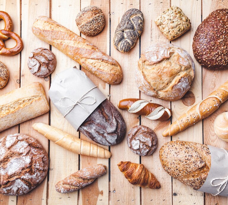 Mehrere Brote und Gebäck auf einem hellen Holztisch. Alles direkt von oben fotografiert. | © iStock/AND-ONE