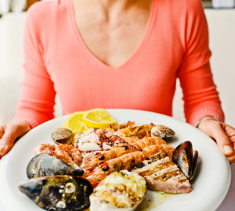 Eine Person im rosa Oberteil hält einen Teller mit Garnelen, Muscheln, Tintenfisch und Zitronenscheiben. | © iStock/anouchka