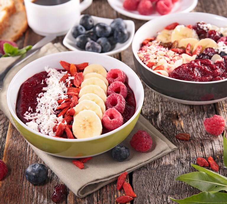 Ein gesundes Frühstück ist auf einem rustikalen Holztisch angerichtet: Zwei Schüsseln mit Acai-Beeren-Smoothie, garniert mit Bananenscheiben, Himbeeren, Kokosraspeln und Goji-Beeren. Daneben sind Schalen mit frischen Blaubeeren und Himbeeren, ein Stück Brot, eine Tasse Kaffee und einige Blätter zu sehen. Der Tisch präsentiert eine reichhaltige Auswahl an farbenfrohen und nahrhaften Lebensmitteln, die eine gesunde Mahlzeit darstellen. | © Getty Images/margouillatphotos