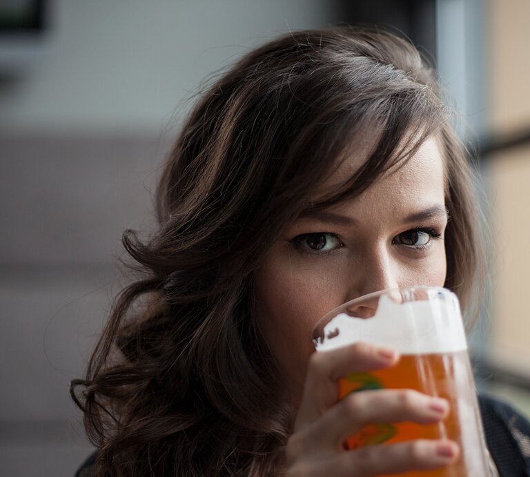 Eine Frau mit lockigem braunem Haar hält ein Glas Bier & trinkt, während sie direkt in die Kamera blickt. | © iStock/brown54486
