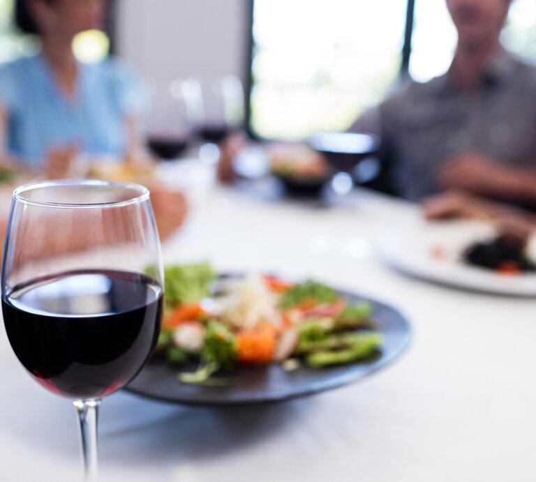 Ein Glas Rotwein im Vordergrund mit scharfem Fokus – im Hintergrund unscharf Personen beim Essen sitzen. | © iStock/Wavebreakmedia
