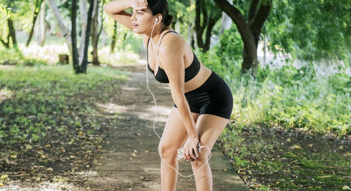 Eine Frau in Sportbekleidung macht eine Pause beim Joggen in einem grünen Park. Sie hält sich die Hand an die Stirn, hat Kopfhörer in den Ohren und sieht erschöpft aus.